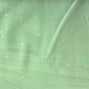 May include: A light green fabric with a subtle texture and small, evenly spaced white dots. The fabric has soft folds and a slightly shiny appearance, suggesting a lightweight material like silk or a blend.
