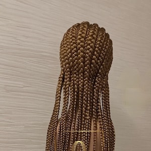 Peut inclure: Une perruque longue tressée de couleur brun clair avec un style droit et fluide. La perruque est sur une tête de mannequin et est coiffée en tresses carrées.
