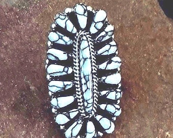 Puede incluir: Un anillo de forma ovalada con una piedra central alargada rodeada por dos filas de piedras más pequeñas en forma de lágrima. Las piedras son de color azul claro con vetas gris oscuro, engastadas en un marco de metal oscuro con un detalle trenzado.