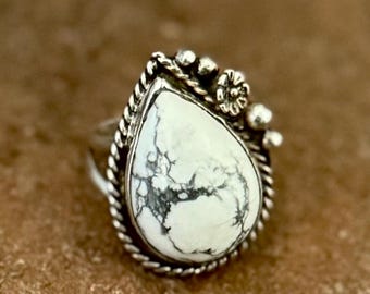 Puede incluir: Un anillo de plata con una piedra blanca en forma de lágrima con vetas gris oscuro. La piedra está engastada en un borde de cuerda de plata trenzada, acentuado con pequeñas esferas de plata y un diminuto detalle floral en un lado.