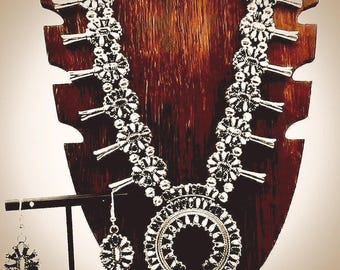 Puede incluir: Un collar tipo squash blossom en tono plateado con múltiples elementos en forma de flor y un colgante central en forma de herradura, acompañado por un par de pendientes colgantes a juego con forma de flor y ganchos plateados.
