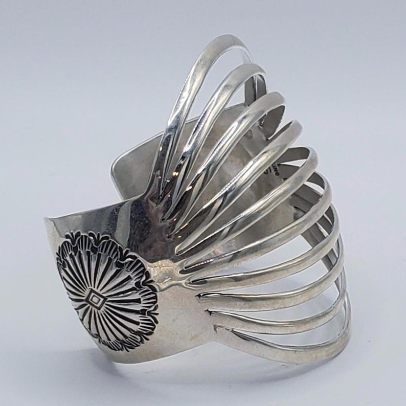 May include: A silver cuff bracelet with a fan-like design. The bracelet features a central floral design with a square center and radiating lines. Multiple curved bars extend from the central design, creating a layered effect. The bracelet has a polished finish.
