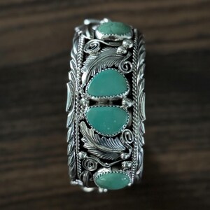 May include: A silver cuff bracelet with turquoise stones. The bracelet features an intricate design with leaf and swirl details. The turquoise stones are oval-shaped and set in the center of the bracelet. The bracelet is displayed on a dark wood surface.
