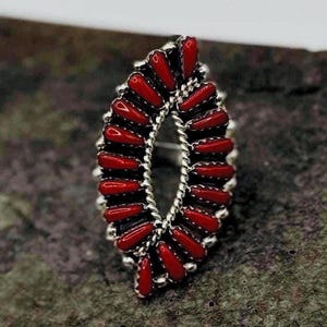May include: A marquise-shaped ring featuring a cluster of small, elongated red stones set in silver. The stones are arranged in a double-row pattern around a central twisted silver wire border, creating a detailed, symmetrical, and elongated design.