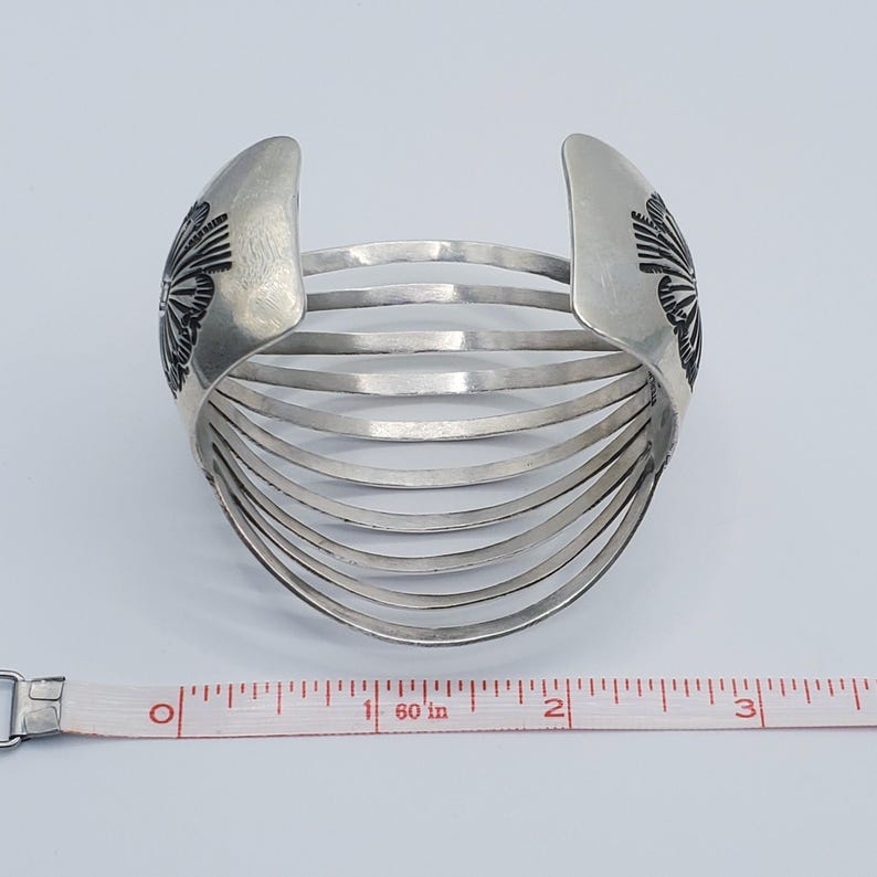 May include: Silver cuff bracelet with a series of parallel bands. The ends of the bracelet feature a stylized floral design. A measuring tape is placed next to the bracelet.