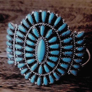 May include: A wide cuff bracelet featuring a large central oval turquoise stone surrounded by multiple concentric rows of smaller, teardrop-shaped turquoise stones set in silver metal with twisted wire detailing and a polished finish.