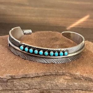 May include: A silver cuff bracelet with a feather design. The bracelet features a row of small, round turquoise stones. The feather design is detailed with etched lines, and the bracelet is displayed on a brown stone surface.