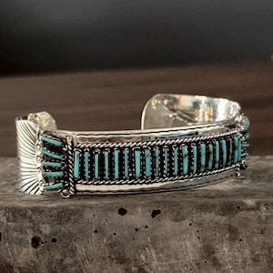 May include: A silver cuff bracelet featuring a central row of vertical, rectangular turquoise inlay stones bordered by twisted silver wire. The ends of the bracelet are decorated with a fan-shaped, etched sunburst pattern in the polished silver metal.