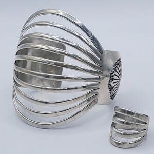 May include: A silver cuff bracelet and ring set. The cuff features a series of curved bars, and the ring has a similar design. The cuff has a decorative floral design on the side. The jewelry is displayed on a white background.