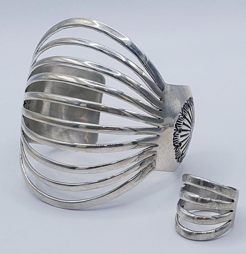 May include: A silver cuff bracelet and ring set. The cuff features a series of curved bars, and the ring has a similar design. The cuff has a decorative floral design on the side. The jewelry is displayed on a white background.