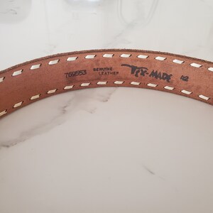 May include: Close-up of a brown leather belt with white stitching. The belt has the text "Genuine Leather" and "Tex-Made" printed on it, along with the number "769553".