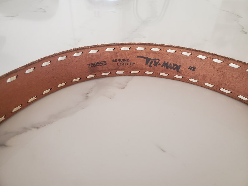 May include: Close-up of a brown leather belt with white stitching. The belt has the text "Genuine Leather" and "Tex-Made" printed on it, along with the number "769553".