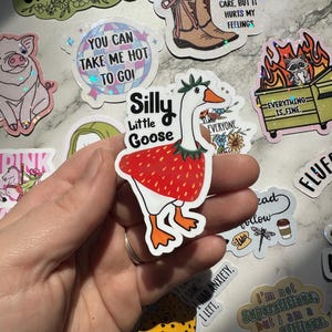 May include: A collection of colorful stickers with various designs. One sticker features a goose dressed as a strawberry with the text "Silly Little Goose." Other stickers include phrases like "You Can Take Me Hot To Go!" and "Everything Is Fine."