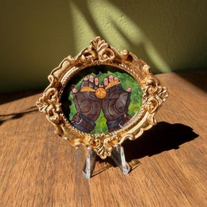 May include: A framed artwork featuring a detailed illustration of two gloved hands holding a golden sphere. The artwork is set within an ornate, gold-colored oval frame, resting on a clear stand, against a green and brown background.