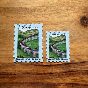 Puede incluir: Dos pegatinas con forma de sello postal azul claro con un diseño de puente de tren escénico. Los sellos presentan un tren rojo que cruza un puente de piedra sobre un valle verde. El texto en los sellos dice "I think we found the train."