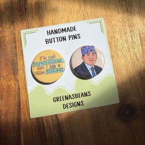 May include: Two handmade button pins on a white card. One pin has the text "I'm not Superstitious, but I am a little Stitious." The other pin features a character wearing a blue bandana. The card reads "GREENASBEANS DESIGNS."