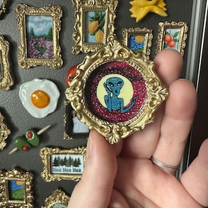 May include: A collection of decorative magnets, including miniature framed art pieces and food-themed designs. One magnet features a gold frame with an alien illustration. Other magnets depict landscapes, fruit, and a fried egg.