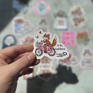 Puede incluir: Una pegatina blanca con un zorro marrón montando una bicicleta rosa con una cesta. El zorro lleva una camisa azul y tiene una cola marrón. La pegatina dice "to the bookstore!" en texto negro.
