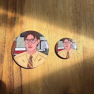 May include: Two circular pin badges with a cartoon illustration of a man wearing glasses, a yellow shirt, and a striped tie. The larger badge is approximately 6.35 cm in diameter, and the smaller one is about 3.81 cm.