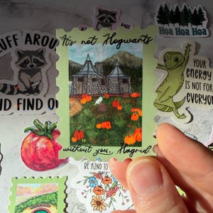 Pode incluir: Um autocolante verde com um design de selo postal com uma ilustração em aquarela de um edifício com abóboras e o texto "It's not Hogwarts without you, Hagrid."