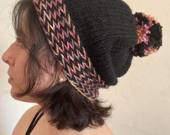 May include: A black knit beanie with a colorful patterned band and a pom-pom. The band features a zig-zag design in shades of pink, yellow, and brown. The pom-pom has similar colors. This winter hat is a cozy accessory.