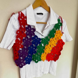 May include: A white short-sleeved shirt with a rainbow crochet top layered over it. The crochet top is made of colourful flowers in red, orange, yellow, green, blue, and purple.