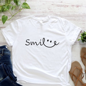May include: White t-shirt with the text "Smile..." in black cursive font, featuring a smiley face with two dots for eyes. The shirt is laid flat on a white wooden surface with a pair of brown sandals, blue jeans, and a small potted plant.