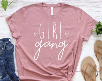 May include: A dusty rose t-shirt with the words "GIRL gang" in white script, with two small heart symbols. The shirt is styled with a knot at the waist, paired with blue jeans and brown sandals. A small plant is also visible.