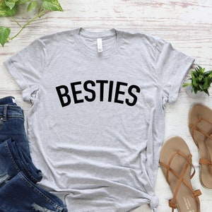 May include: A gray t-shirt with the word "BESTIES" printed in black on the front. The shirt is laid flat on a white wooden surface with a pair of blue jeans, a small potted plant, and a pair of brown sandals.