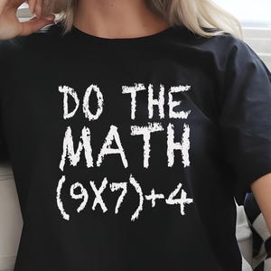 May include: Black t-shirt with the phrase "DO THE MATH (9X7)+4" printed in white. The shirt is worn with blue jeans. The text is in a bold, handwritten style.