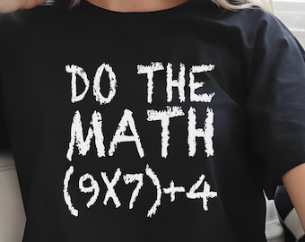 May include: Black t-shirt with the phrase "DO THE MATH (9X7)+4" printed in white. The shirt is worn with blue jeans. The text is in a bold, handwritten style.