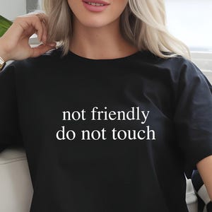 May include: Black t-shirt with the white text "not friendly do not touch." The shirt is worn with light blue jeans. The model is wearing gold bracelets.