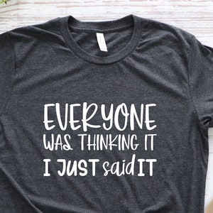May include: Dark gray t-shirt with the text "EVERYONE WAS THINKING IT I JUST SAID IT" in white. The shirt is paired with blue jeans and brown sandals. A small green plant is also visible.
