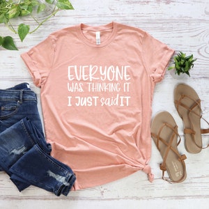 May include: A peach-colored t-shirt with the text "EVERYONE WAS THINKING IT I JUST SAID IT" in white. The shirt is paired with blue denim jeans and tan sandals. Green plants are arranged around the clothing.