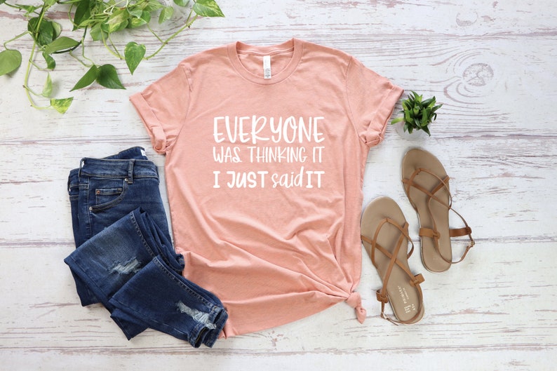 May include: A peach-colored t-shirt with the text "EVERYONE WAS THINKING IT I JUST SAID IT" in white. The shirt is paired with blue denim jeans and tan sandals. Green plants are arranged around the clothing.