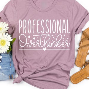 a purple t-shirt with the text "Professional Overthinker" printed on it, along with a pair of sandals and some flowers.