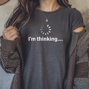 May include: Dark gray t-shirt with the text "I'm thinking...." and a loading circle graphic. The shirt is paired with a patterned jacket and a necklace. The overall style is casual and comfortable.