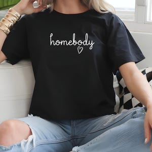 Puede incluir: Camiseta negra con la palabra "homebody" en escritura blanca y un peque&ntilde;o coraz&oacute;n. La camiseta se combina con vaqueros azul claro. La est&eacute;tica general es informal y c&oacute;moda.