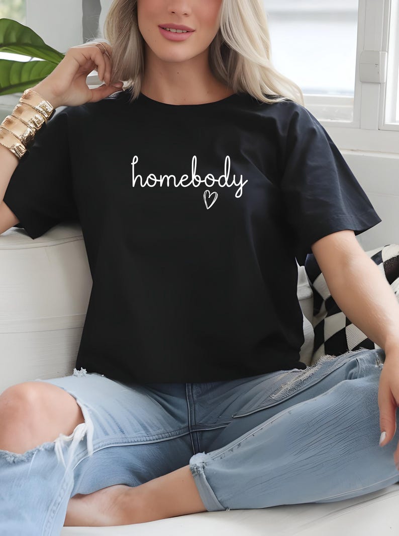 Puede incluir: Camiseta negra con la palabra "homebody" en escritura blanca y un peque&ntilde;o coraz&oacute;n. La camiseta se combina con vaqueros azul claro. La est&eacute;tica general es informal y c&oacute;moda.