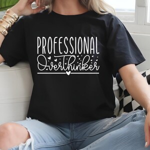 A woman wearing a black t-shirt with white text that says "Professional Overthinker" is sitting on a couch.