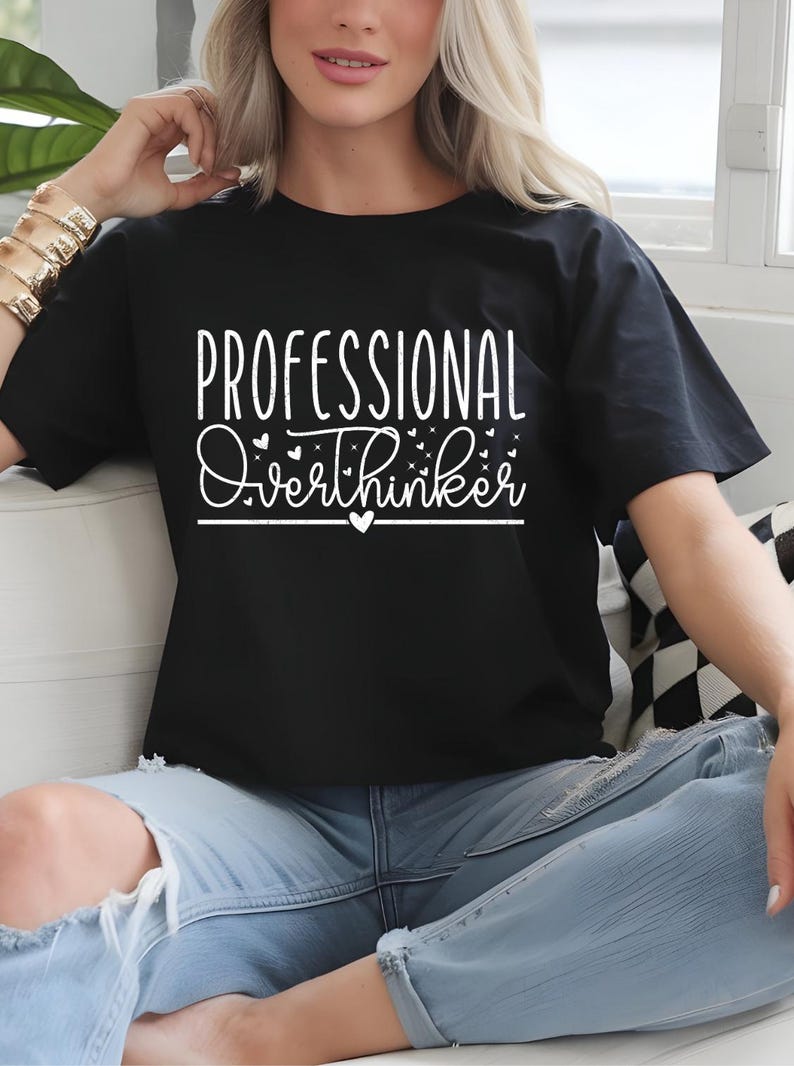 A woman wearing a black t-shirt with white text that says "Professional Overthinker" is sitting on a couch.