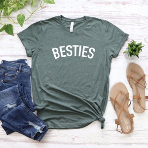 May include: A green t-shirt with the word "BESTIES" printed in white on the front. The shirt is laid flat on a white wooden surface with a pair of brown sandals and a pair of blue jeans.