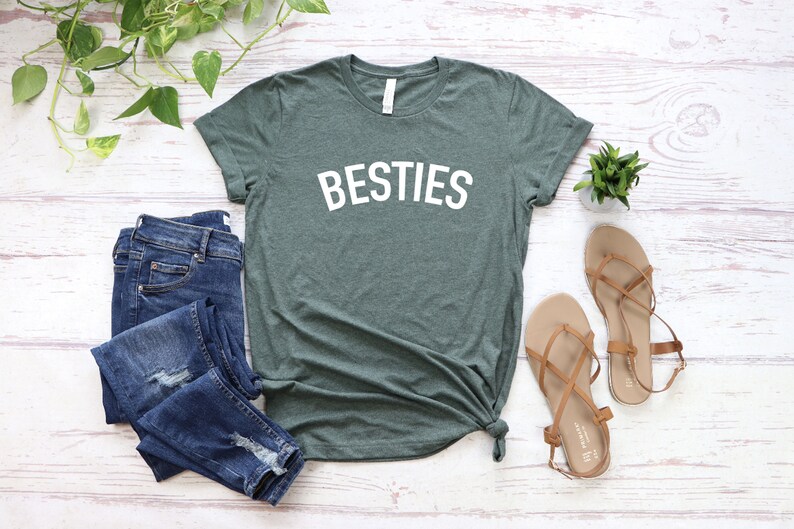 May include: A green t-shirt with the word "BESTIES" printed in white on the front. The shirt is laid flat on a white wooden surface with a pair of brown sandals and a pair of blue jeans.