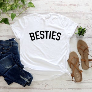 May include: White t-shirt with the word "BESTIES" printed in black on the front. The shirt is laid flat on a white wooden surface with a pair of brown sandals, blue jeans, and a small potted plant.