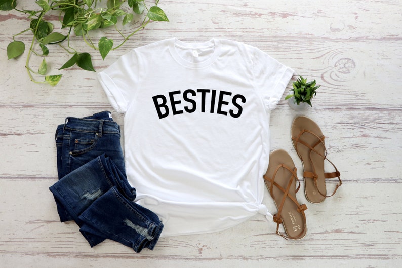 May include: White t-shirt with the word "BESTIES" printed in black on the front. The shirt is laid flat on a white wooden surface with a pair of brown sandals, blue jeans, and a small potted plant.