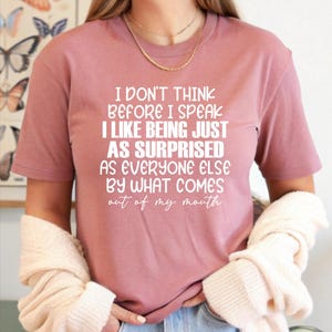 May include: Dusty rose t-shirt with white text: "I DON'T THINK BEFORE I SPEAK I LIKE BEING JUST AS SURPRISED AS EVERYONE ELSE BY WHAT COMES out of my mouth." The shirt is paired with blue jeans. A cream-colored sweater is draped over the arms.