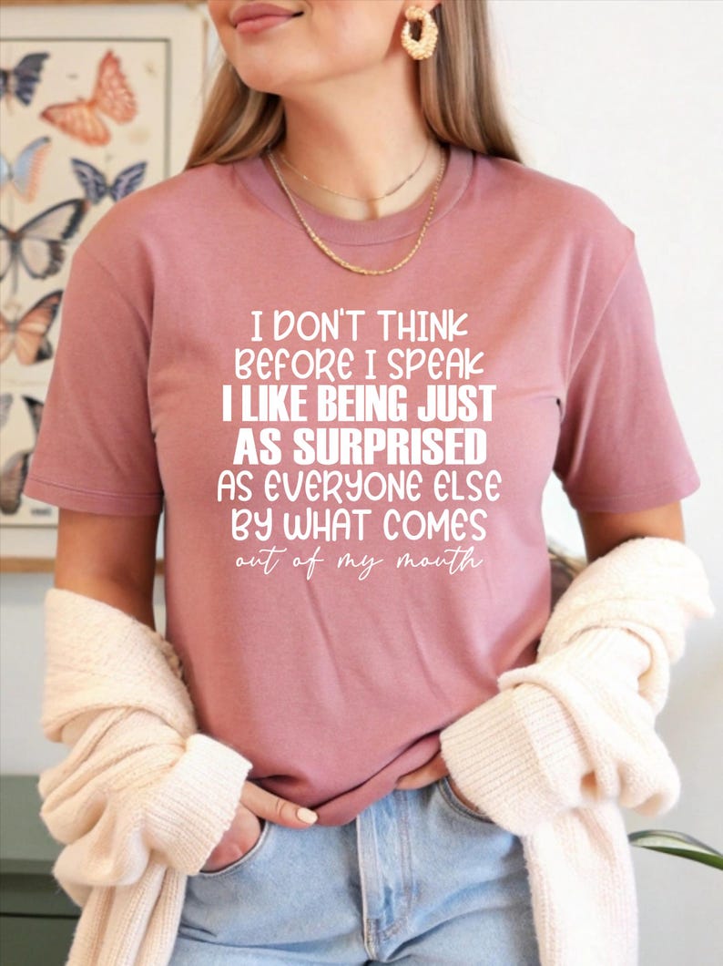 May include: Dusty rose t-shirt with white text: "I DON'T THINK BEFORE I SPEAK I LIKE BEING JUST AS SURPRISED AS EVERYONE ELSE BY WHAT COMES out of my mouth." The shirt is paired with blue jeans. A cream-colored sweater is draped over the arms.