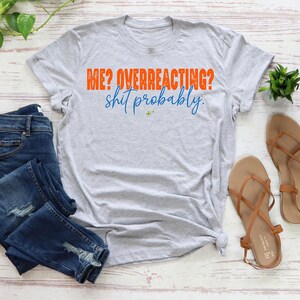 May include: A light gray short-sleeved t-shirt featuring the orange text ME? OVERREACTING? and the blue cursive text shit probably. The shirt is displayed alongside dark blue distressed denim jeans and a pair of tan strappy sandals.
