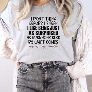 May include: A light gray t-shirt with the text "I DON'T THINK BEFORE I SPEAK I LIKE BEING JUST AS SURPRISED AS EVERYONE ELSE BY WHAT COMES out of my mouth." The shirt is paired with gray acid wash jeans.