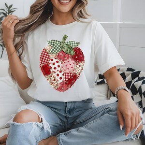 May include: A white short-sleeved t-shirt featuring a large, central strawberry graphic composed of various red, white, and green fabric-style patches. The strawberry design includes patterns like gingham, small hearts, and floral prints with a green leaf top.